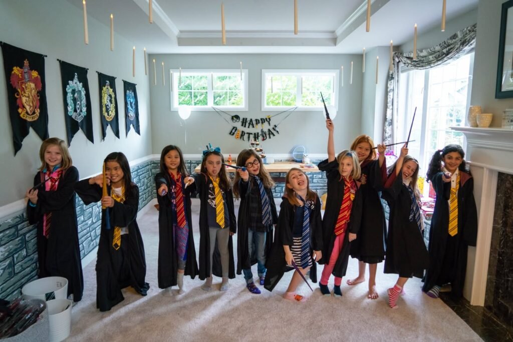 A group of children dress in wizard hats, wands, and scarves at a Harry Potter birthday party. The scene is decorated with house banners and a happy birthday garland.