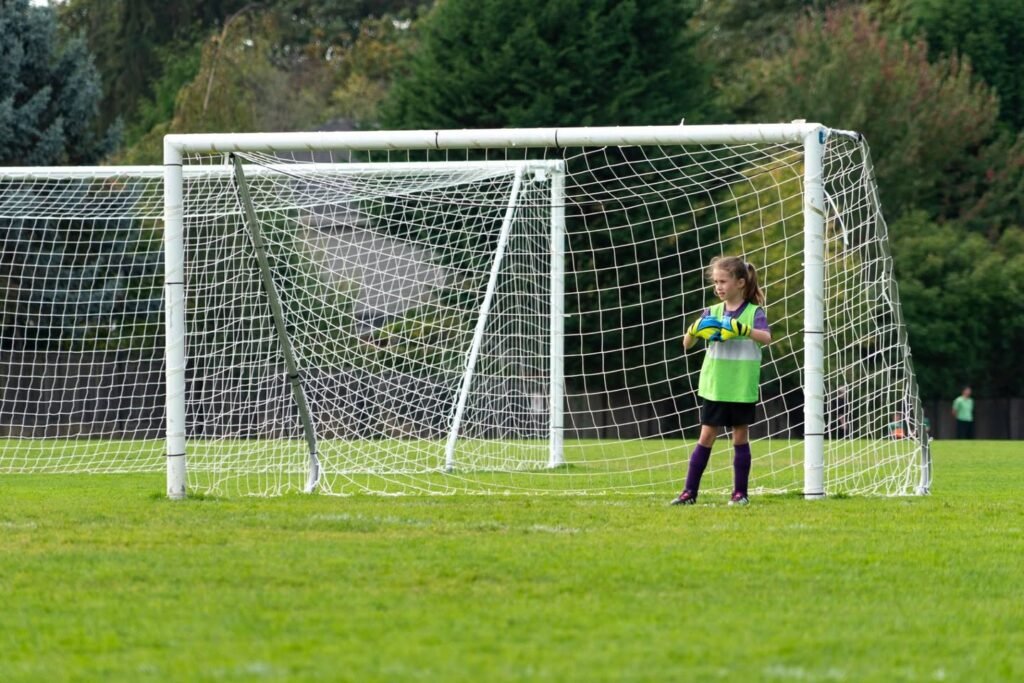 A young goalkeeper in a bright green shirt stands ready in front of the soccer goal on a grassy field.