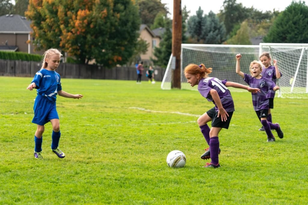 A young player in blue defends the ball near the goal. Teammates in purple jerseys celebrate nearby on the grassy field.