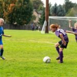 A young player in blue defends the ball near the goal. Teammates in purple jerseys celebrate nearby on the grassy field.
