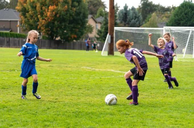 A young player in blue defends the ball near the goal. Teammates in purple jerseys celebrate nearby on the grassy field.