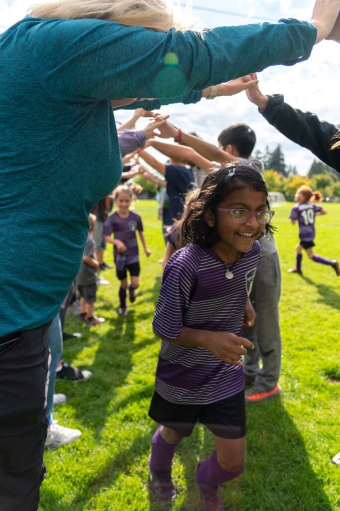 A young soccer player smiles while teammates form a lineup to give high-fives on a sunny day.