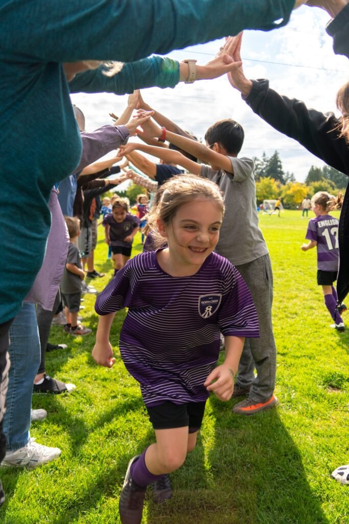 A youth soccer player in a purple uniform runs through a celebratory tunnel formed by teammates and fans.