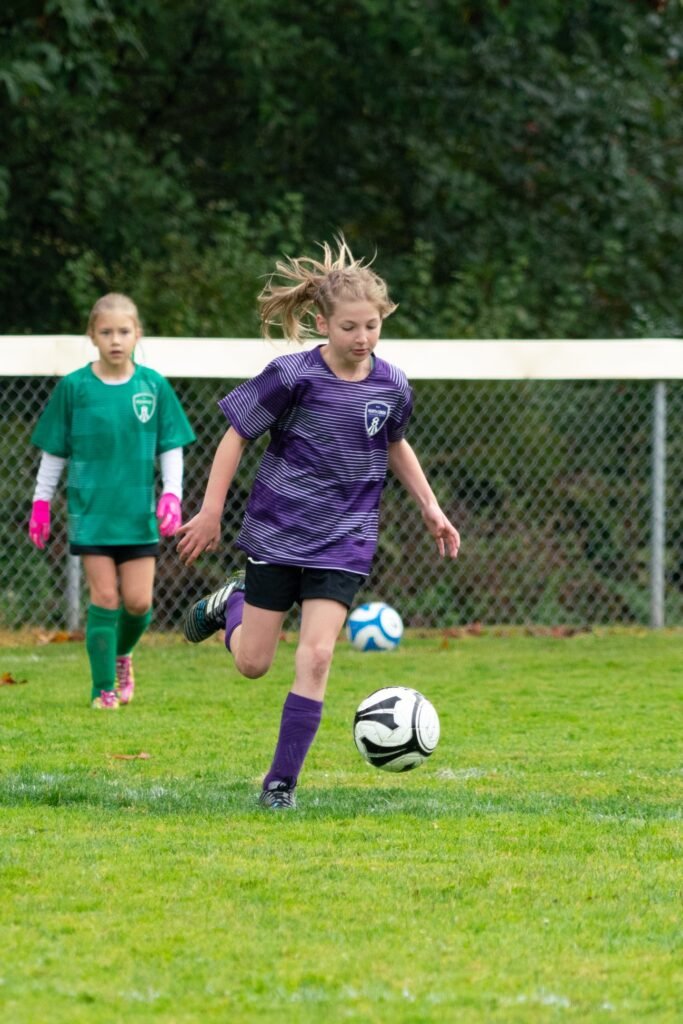 A young player in a purple jersey chases a soccer ball on a green field during a game.