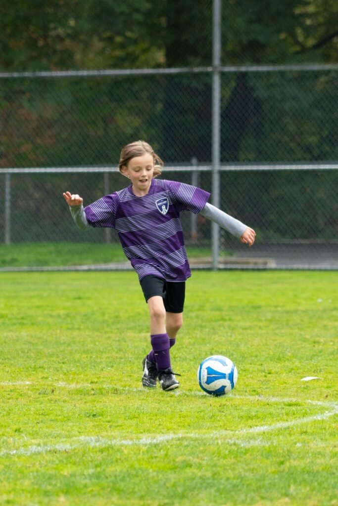 A young soccer player in a purple jersey kicks a ball on a grassy field in the Pacific Northwest.