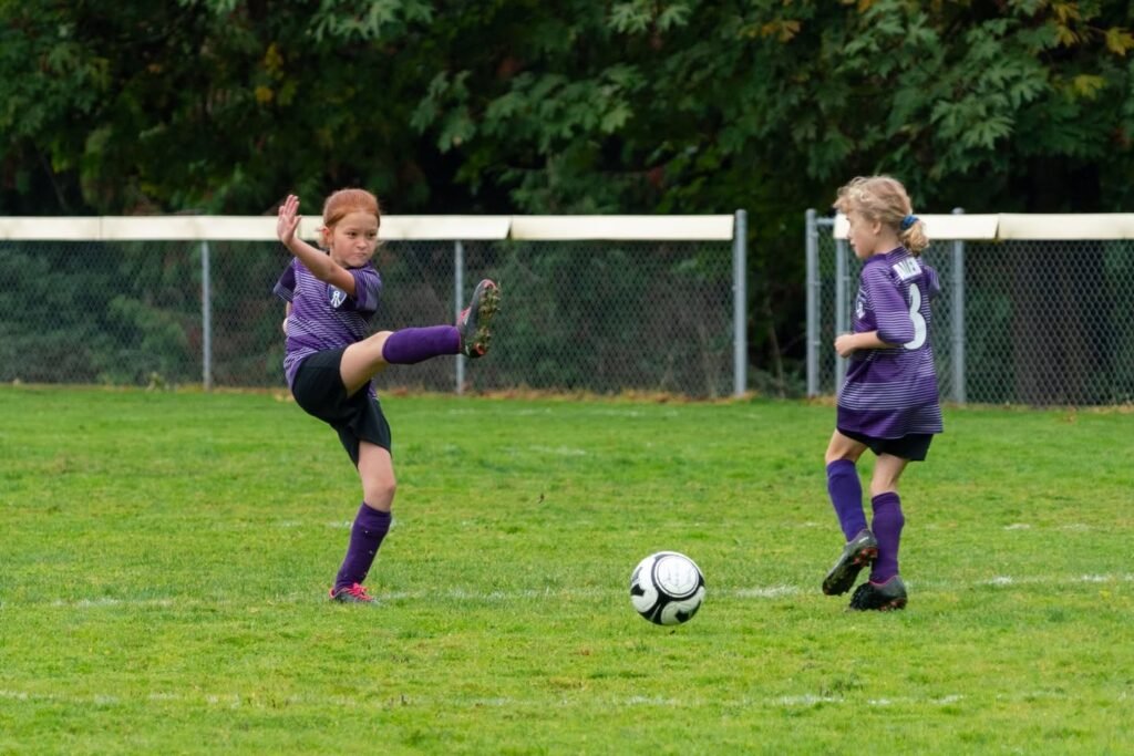 A young athlete in a purple jersey kicks a soccer ball. A teammate in matching gear watches closely on the grassy field.