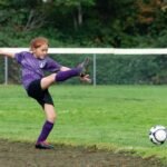 A soccer player in a purple uniform kicks a ball during an outdoor game.