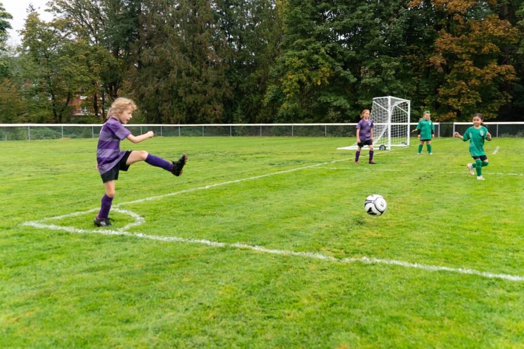 A youth soccer game in progress on a grassy field with a soccer player mid-kick and a ball near the center circle.