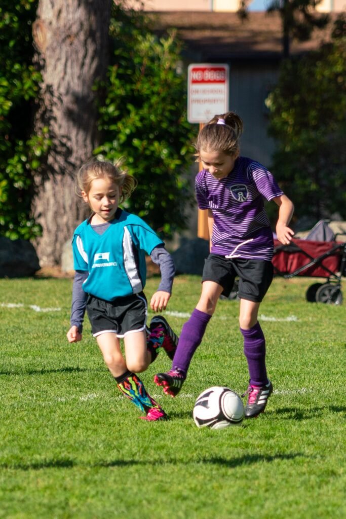 Two kids in soccer uniforms actively play on a green grass field near a tree.