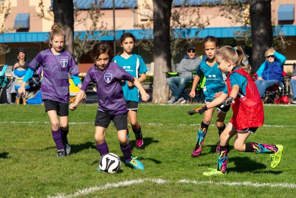 A youth soccer match in progress with kids in purple and teal jerseys competing on a sunny grass field.