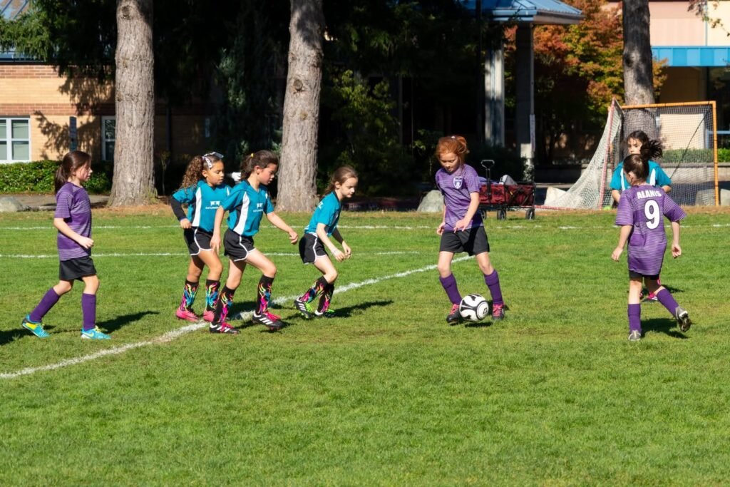 Children in purple and turquoise jerseys playing soccer on a green field with trees and a building in the background.