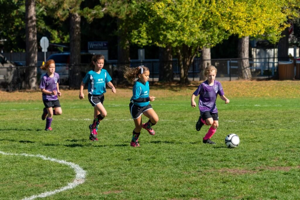 Young soccer players in teal and purple uniforms chase the ball on a grassy field. Trees and a fence are visible in the background.