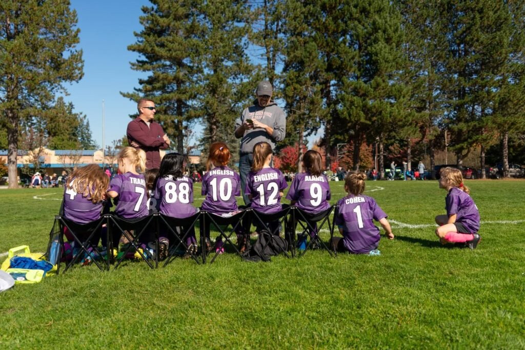 Children in matching purple sports jerseys sit on a grassy field framed by tall evergreen trees under a clear blue sky.