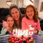 A family gathers around a birthday cake with candles. A woman and two girls smile together inside a home.