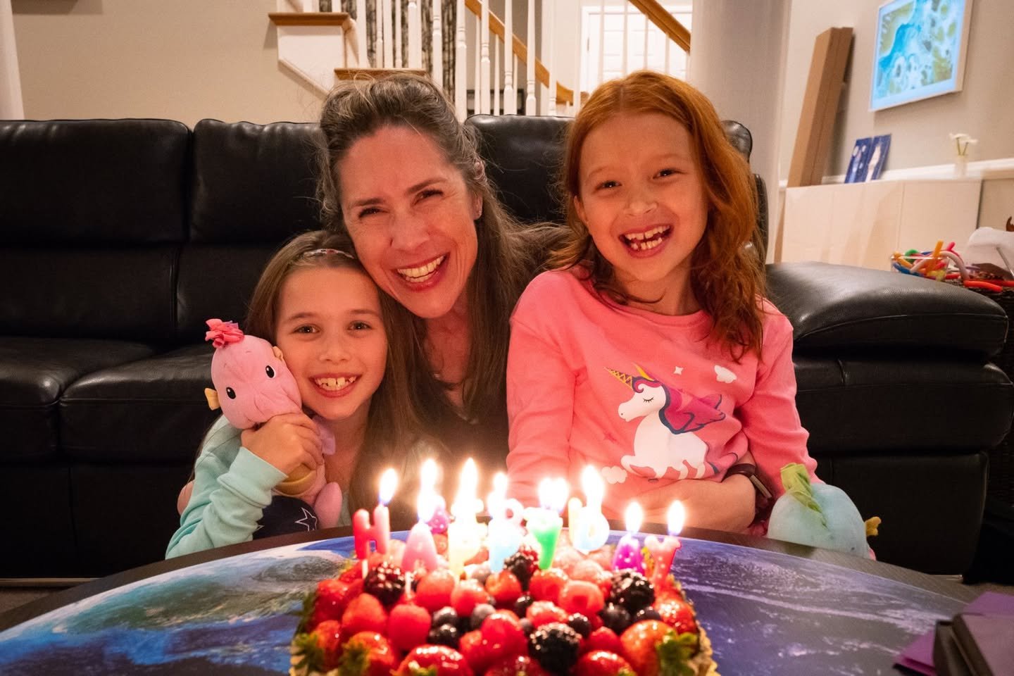 A family gathers around a birthday cake with candles. A woman and two girls smile together inside a home.