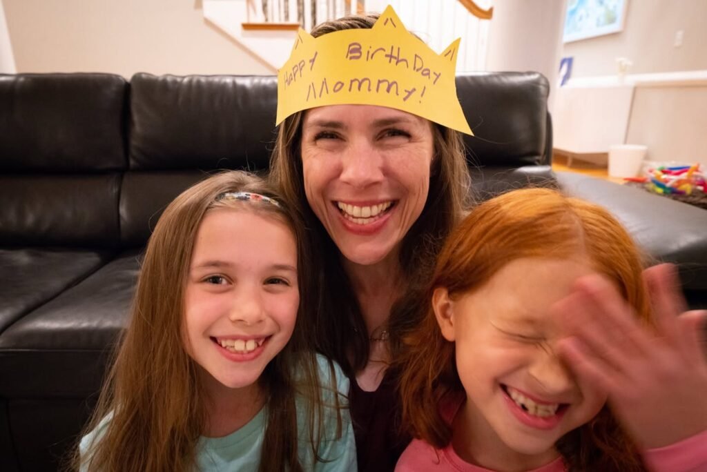 A joyful family celebrating a birthday with a handmade paper crown on the couch. The woman in the middle wears a crown that reads 'Happy Birthday Mommy.'