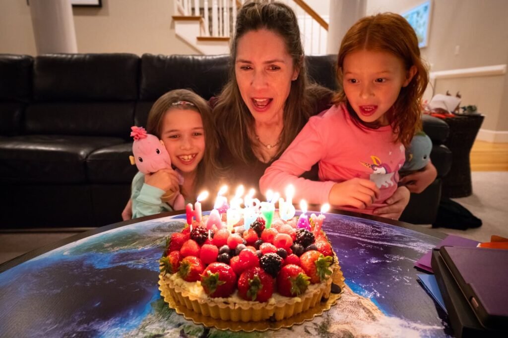A family gathers around a table to celebrate, with a beautiful fruit tart with lit candles in the center.