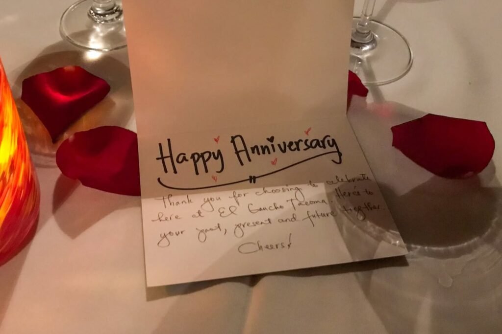 A written 'Happy Anniversary' card on a table surrounded by red rose petals.