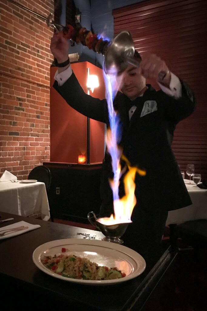A server performs a fiery tableside dessert presentation with vibrant blue and orange flames rising from a metal pan next to a plated dish.