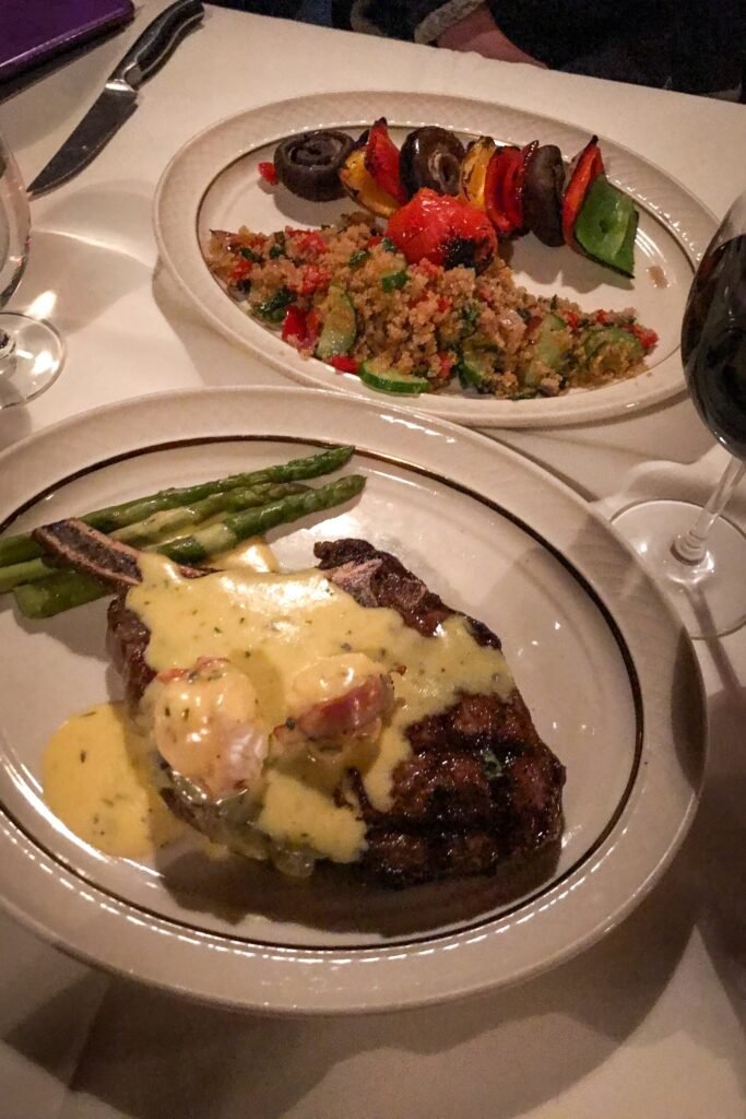 A plated steak and cream sauce with asparagus served alongside a salad and roasted vegetables. A glass of red wine and knife rest on the table for a cozy dinner.