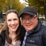 A selfie of two smiling people in jackets and a cap, with autumn-colored trees and a railing in the background.