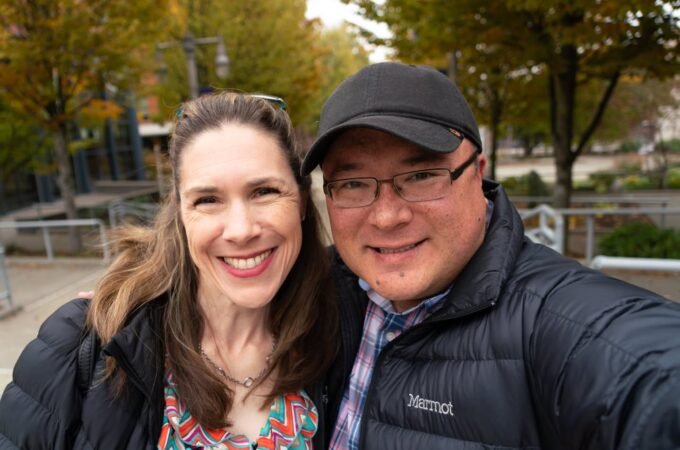 A selfie of two smiling people in jackets and a cap, with autumn-colored trees and a railing in the background.