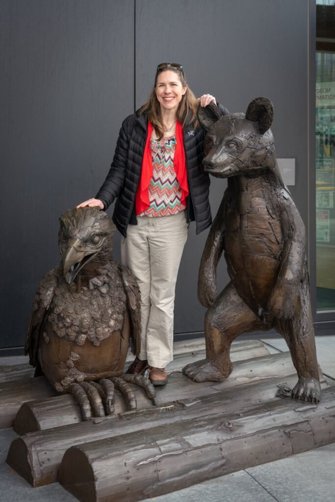 A traveler poses next to bronze sculptures of a bird and a bear in an urban setting. The detailed statues are based on wooden log structures.
