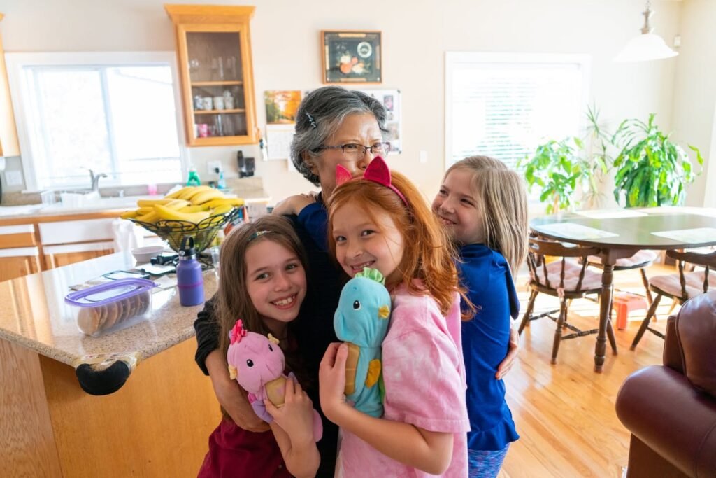 A grandmother warmly hugs her three young granddaughters in a bright home kitchen.