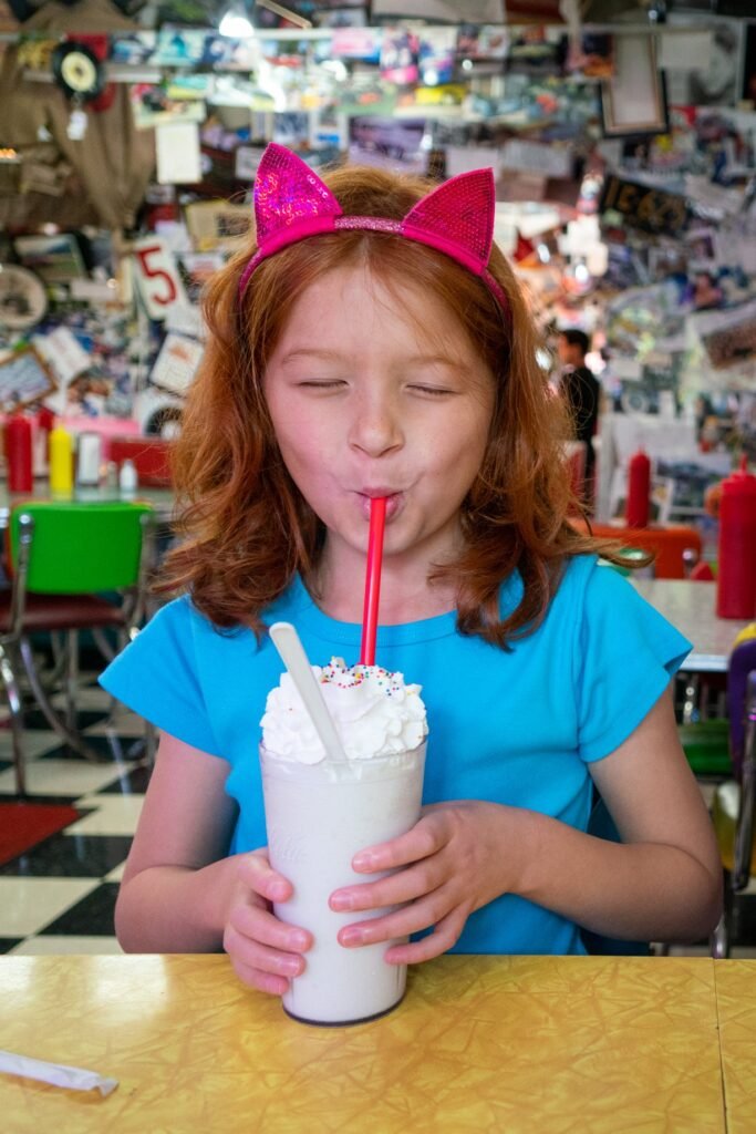 A child in a blue shirt and pink glittery cat ears enjoys a milkshake in a diner. The background features a wall covered in postcards.