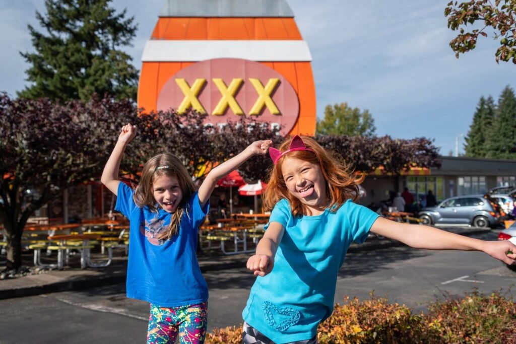 Children smiling with spread arms in front of a bright, retro-style XXX sign