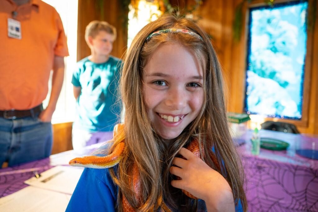 A child with long hair smiles at the camera at a community event. In the background, other children and an adult in an orange shirt stand near a table with purple cloth.