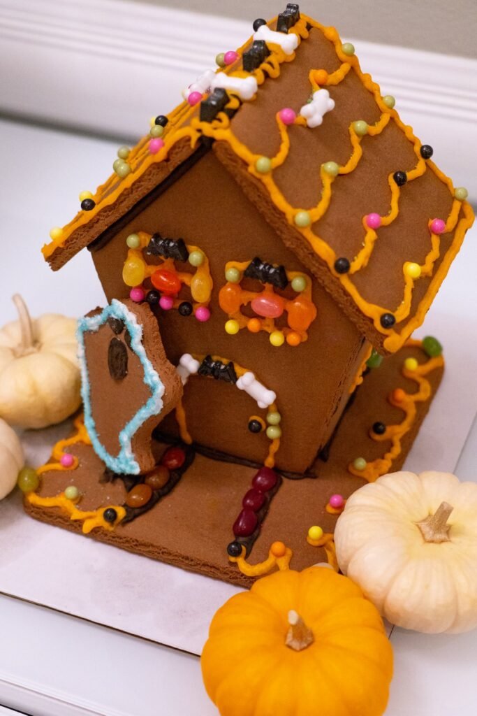 A decorated gingerbread house surrounded by small pumpkins, showcasing intricate details and a festive autumn theme. The house is adorned with colorful candies and icing, set on a rustic wooden surface.