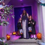 Cannon Beach Thanksgiving Halloween porch decorated with lit jack-o-lanterns, an '800' sign, and a starry moon overlay. Two children holding collection buckets pose with an adult under a glowing purple light.