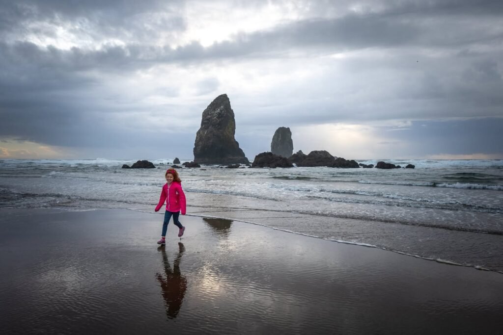 A person in a pink jacket walking on a wet beach with mossy sea stacks in the background. The overcast sky reflects on the sand surface.
