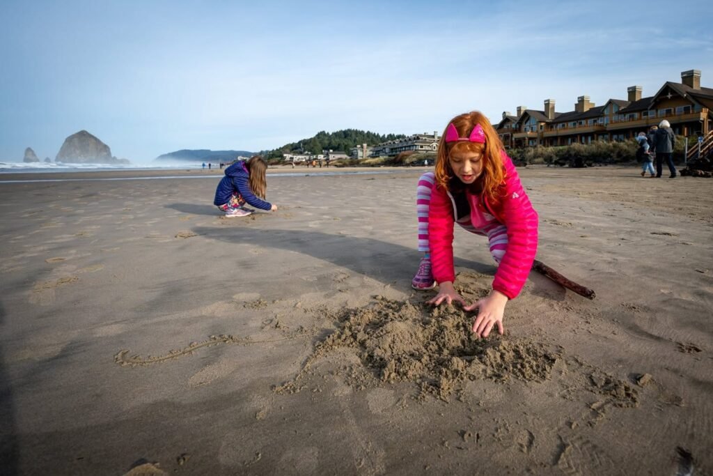 Two people enjoy a day of sand play on the beach at Cannon Beach, Oregon, with the iconic Haystack Rock in the distance.