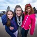 A woman and two girls pose together on a sandy Pacific Northwest beach with coastal rocks and a cloudy sky in the background. They are wearing colorful winter jackets and smiling.