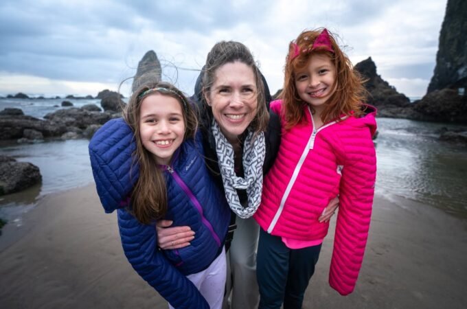 A woman and two girls pose together on a sandy Pacific Northwest beach with coastal rocks and a cloudy sky in the background. They are wearing colorful winter jackets and smiling.