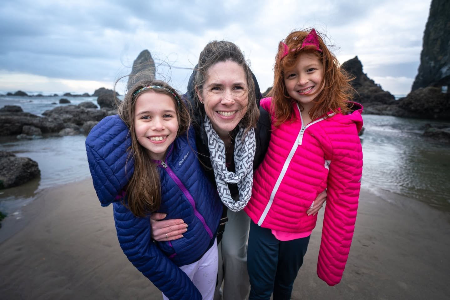 Cannon Beach Thanksgiving A woman and two girls pose together on a sandy Pacific Northwest beach with coastal rocks and a cloudy sky in the background. They are wearing colorful winter jackets and smiling.