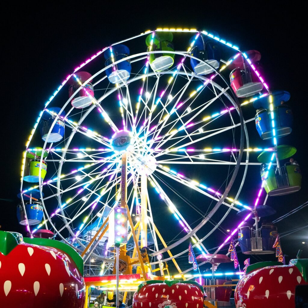 A vibrant Ferris wheel decorated with multi-colored lights against a dark night sky, with strawberry-shaped decorations in the foreground.