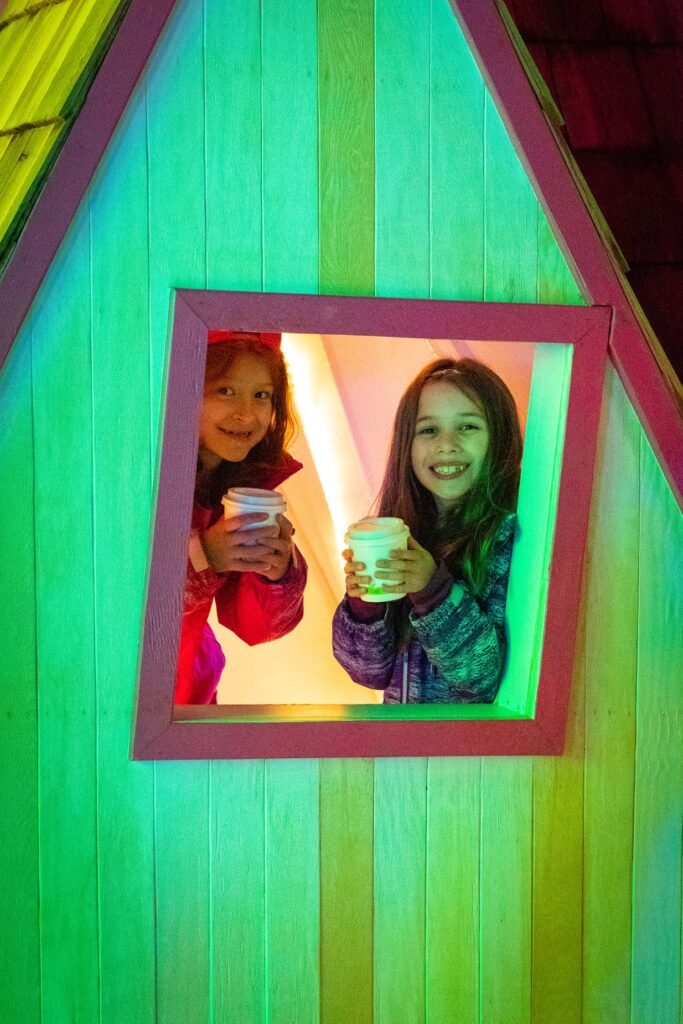 Two children smile from a colorful Toyland house window while holding drinks, illuminated by vibrant green and yellow lighting.