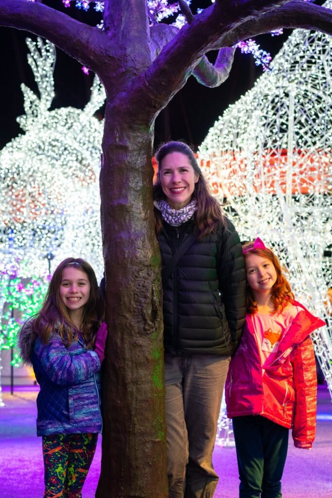 Three people stand together against a tree with colorful holiday light decorations in the background.