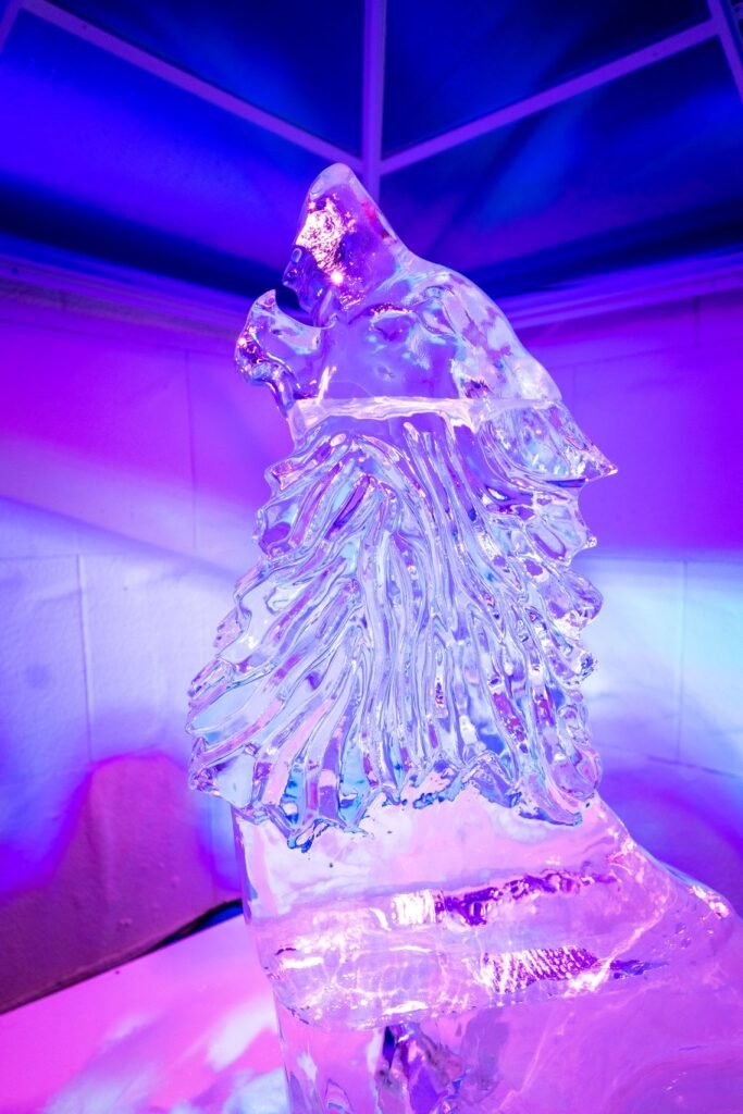 A detailed ice sculpture of a human figure carved in a flowing robe, illuminated by vibrant purple and blue lighting.