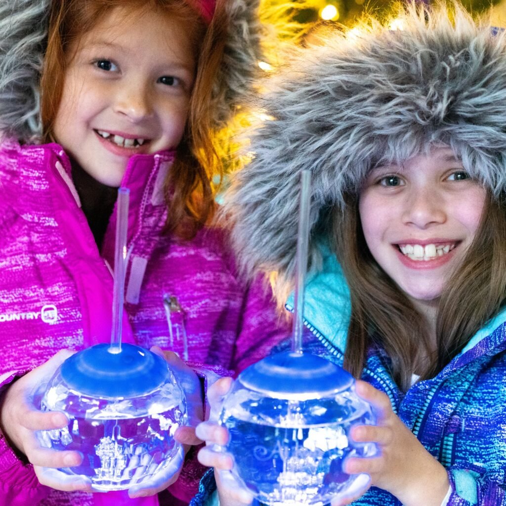 Two smiling children wearing fluffy winter hats hold clear novelty mugs with straws, bundled up in colorful jackets. Festive lights twinkle softly in the background.