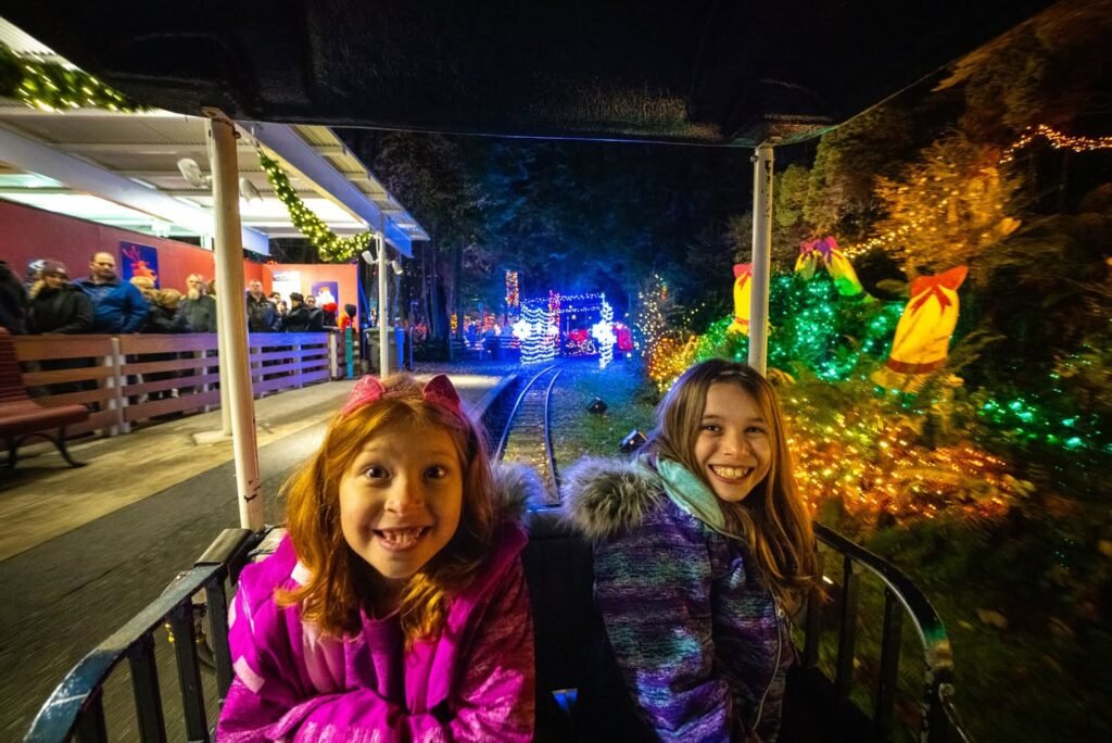 Two smiling girls in winter jackets seated on a ride train at a nighttime Christmas light display. The track is surrounded by colorful illuminated decorations, holiday ornaments, and festive greenery.