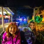 Two smiling girls in winter jackets seated on a ride train at a nighttime Christmas light display. The track is surrounded by colorful illuminated decorations, holiday ornaments, and festive greenery.