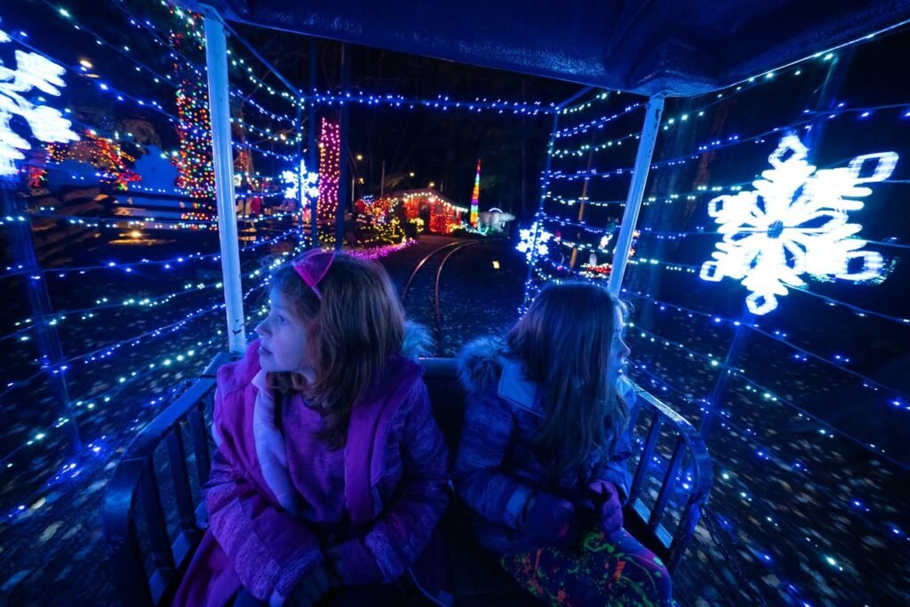Two people in winter coats sit side by side in a seat, looking out through a vehicle window onto a tunnel of blue and white Christmas lights with large glowing snowflakes.
