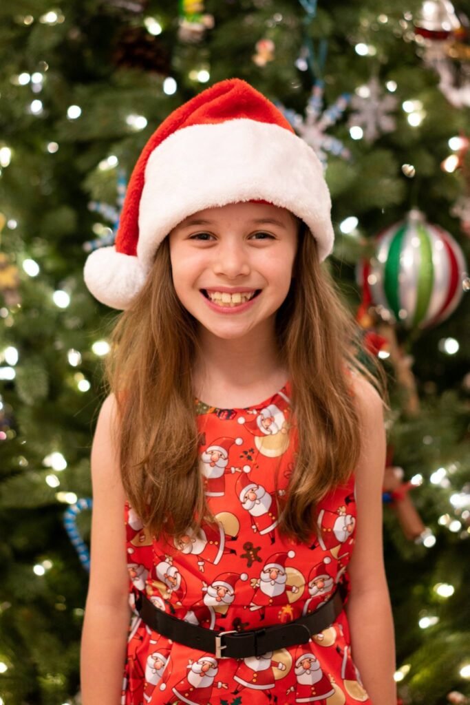 A joyful individual wearing a Santa hat and festive red dress smiles in front of a decorated Christmas tree.