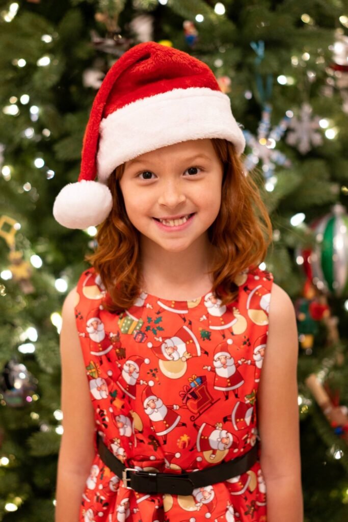 Smiling child wearing a Santa hat and a red patterned dress standing in front of a decorated Christmas tree.