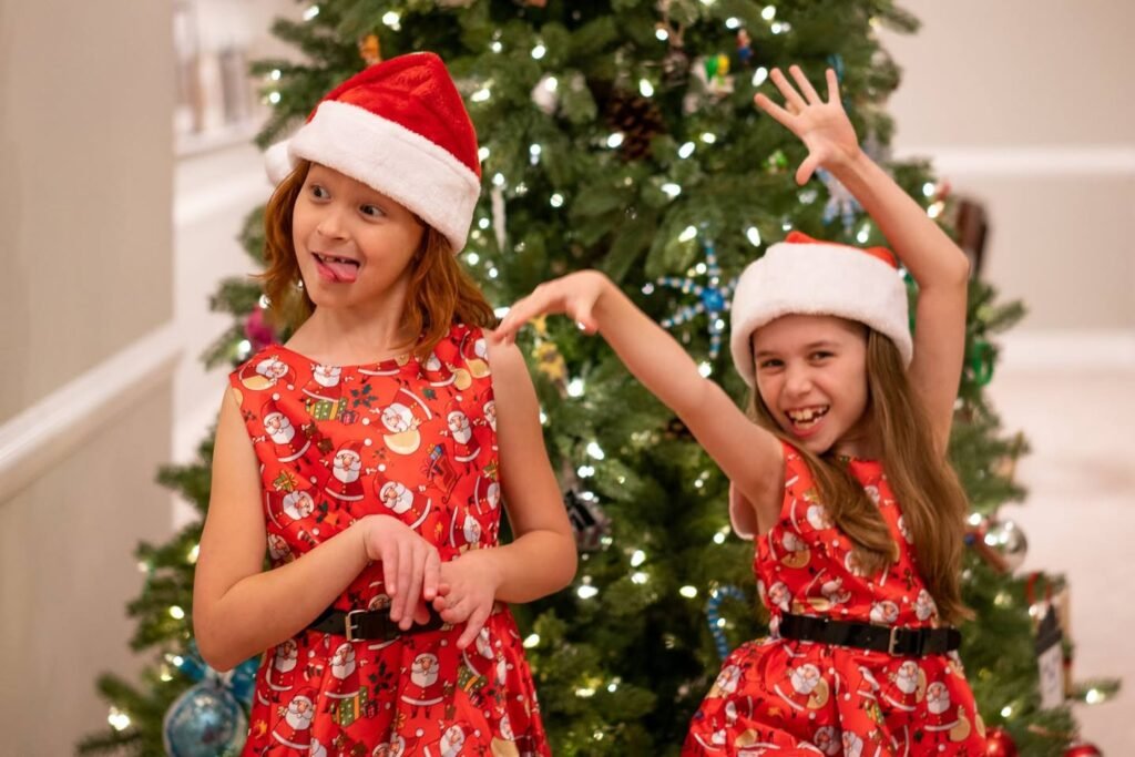 Two girls in red Santa-themed dresses and hats stand in front of a decorated Christmas tree.