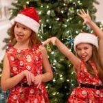 Two girls in red Santa-themed dresses and hats stand in front of a decorated Christmas tree.